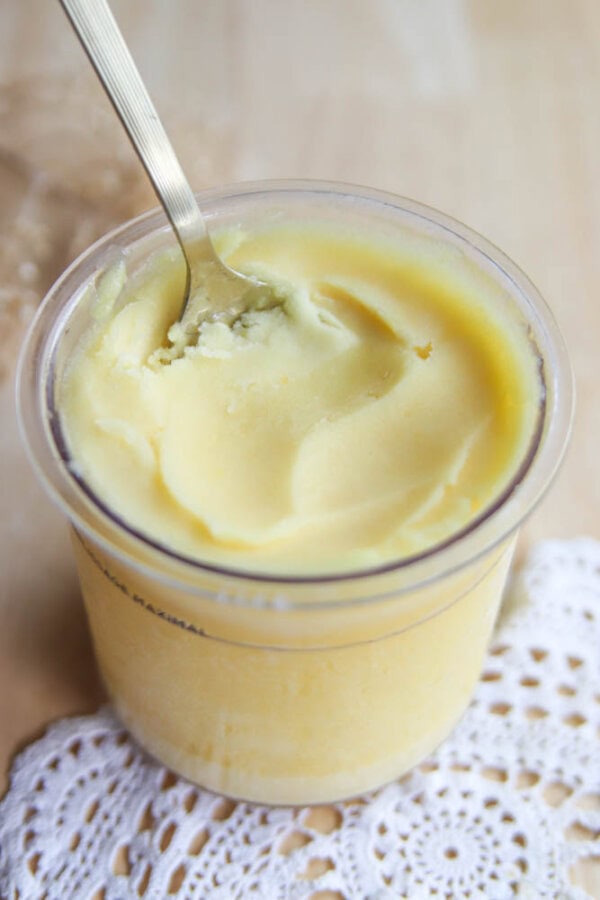 A spoon rests in a clear jar filled with smooth, yellow ghee—reminiscent of the vibrant hues of ninja creami peach sorbet—placed on a crocheted white doily on a wooden surface.