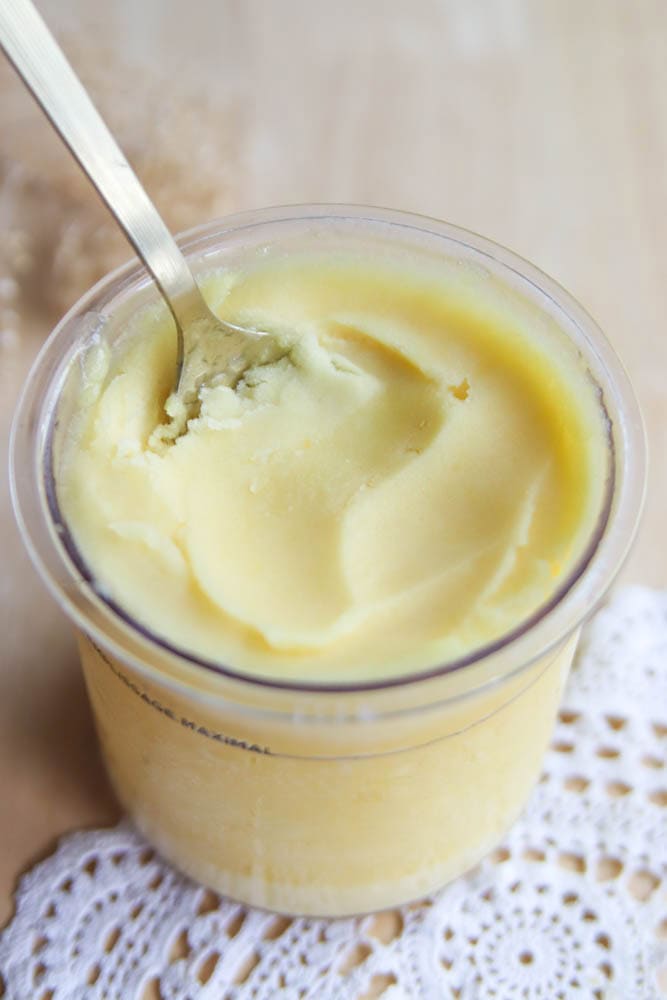 A spoon scooping creamy, yellow butter from a clear plastic container, placed on a beige surface with part of a white lace doily visible underneath—reminiscent of the silky texture found in Ninja Creami peach sorbet.