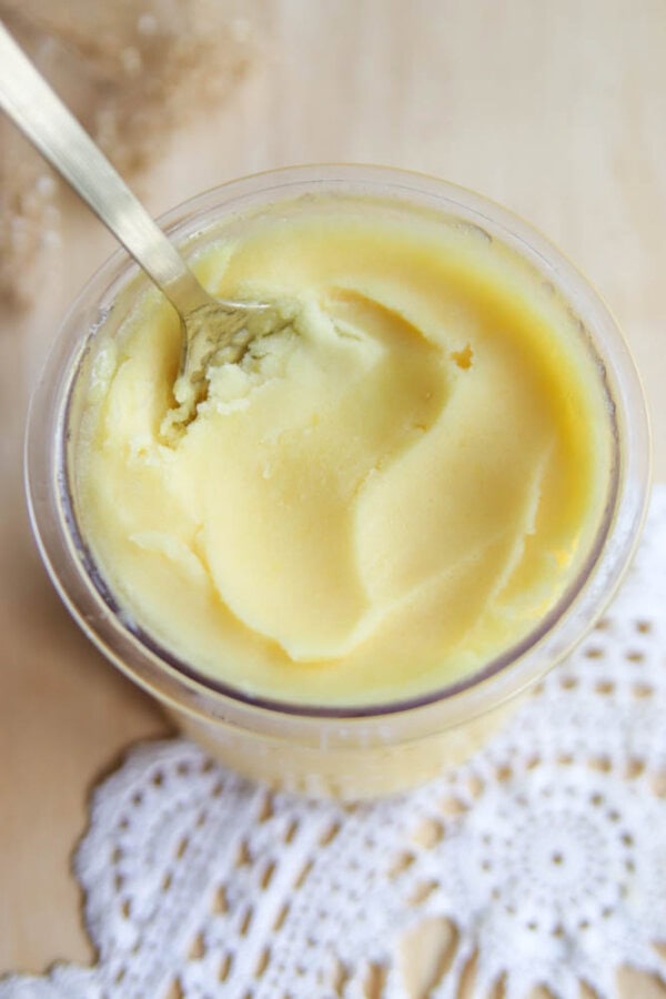 A top-down view of a jar filled with creamy yellow body butter, reminiscent of ninja creami peach sorbet, and a spoon scooping some out. The jar sits on a white lace doily atop a light wooden surface.