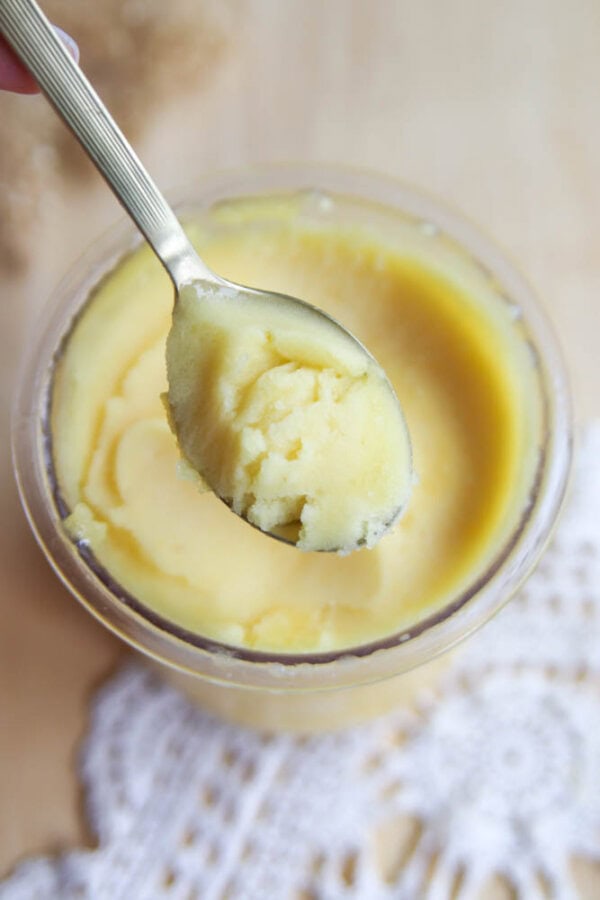 A silver spoon holds a scoop of yellow ghee above a glass jar filled with more ghee, resting on a light wooden surface with a lace doily partially visible, resembling the smooth texture of Ninja Creami peach sorbet.