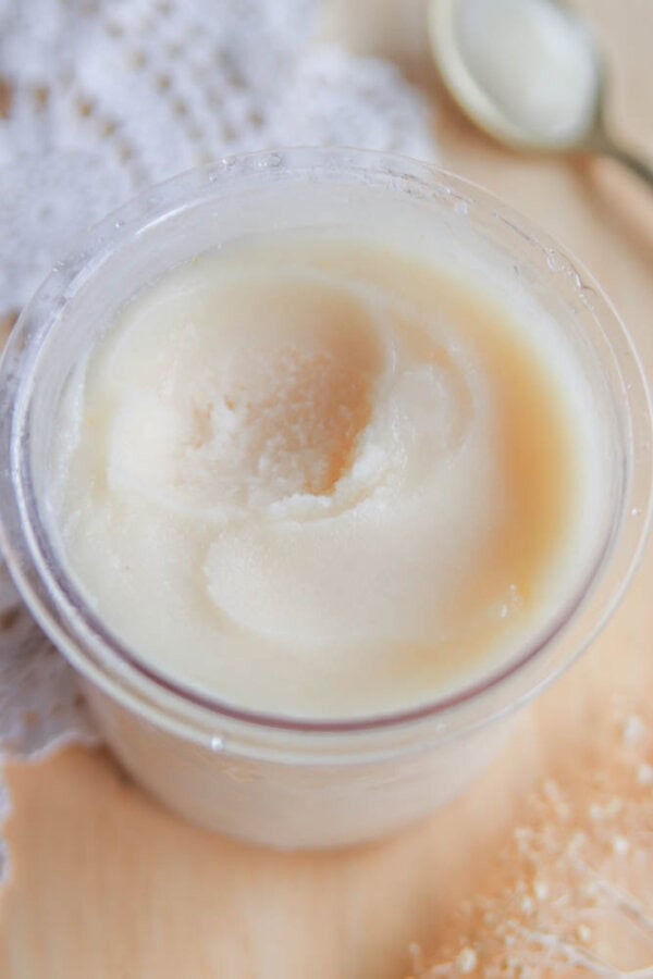A clear container filled with a creamy, off-white substance, resembling a luscious body butter or balm, sits on a wooden surfaceโwith a lace cloth and a spoon in the backgroundโevoking the smooth texture of a ninja creami pear sorbet recipe.