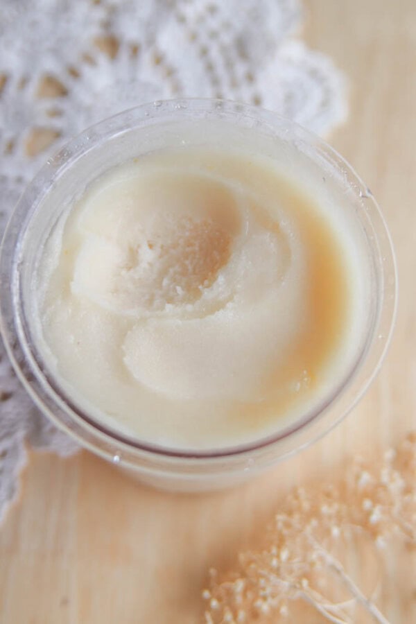 An open jar of creamy, pale yellow body butter sits on a wooden surface, near a white lace cloth and some dried flowersโits smooth texture reminiscent of a freshly made ninja creami pear sorbet recipe.