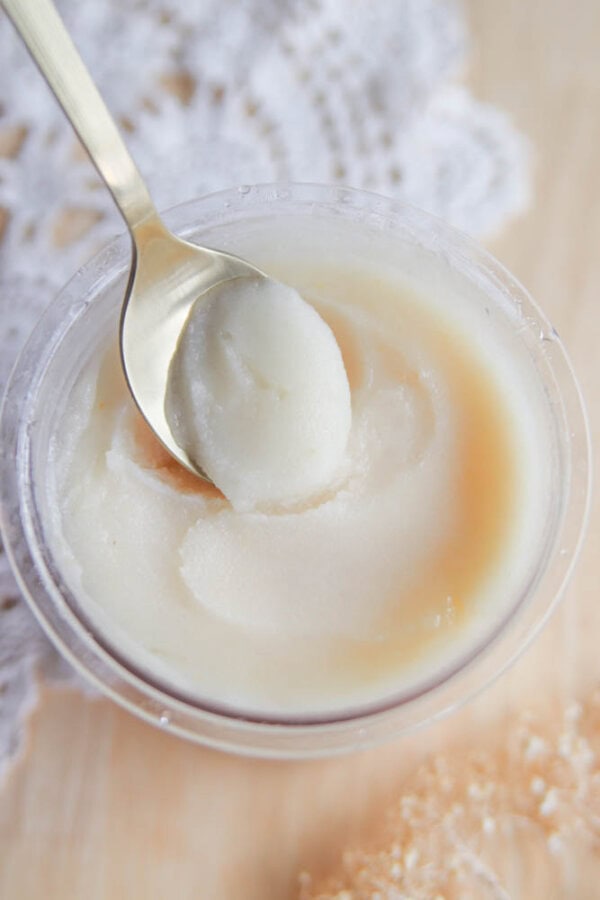 A close-up of a spoon scooping out a creamy white treat, possibly inspired by a Ninja Creami pear sorbet recipe, from a small clear container placed on a light surface with lacy fabric in the background.