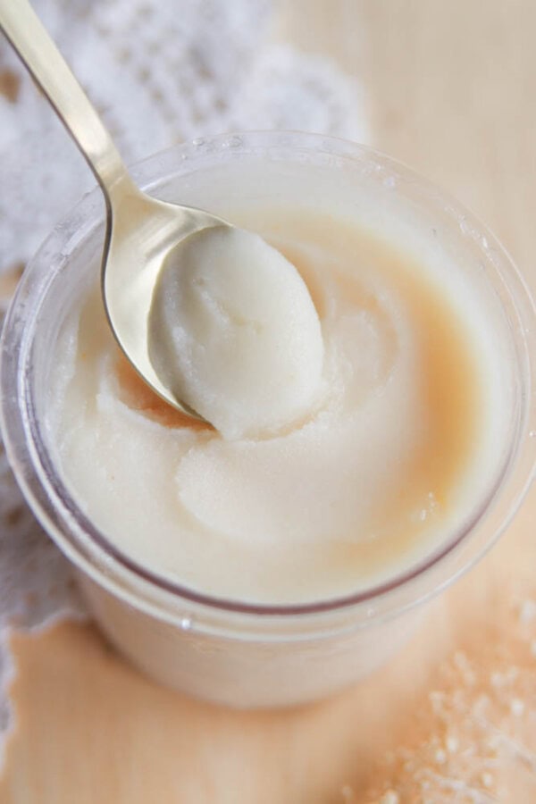 A close-up of a spoon scooping a creamy, white substanceโresembling a treat from a Ninja Creami pear sorbet recipeโfrom a clear plastic container on a light wooden surface with lace cloth in the background.