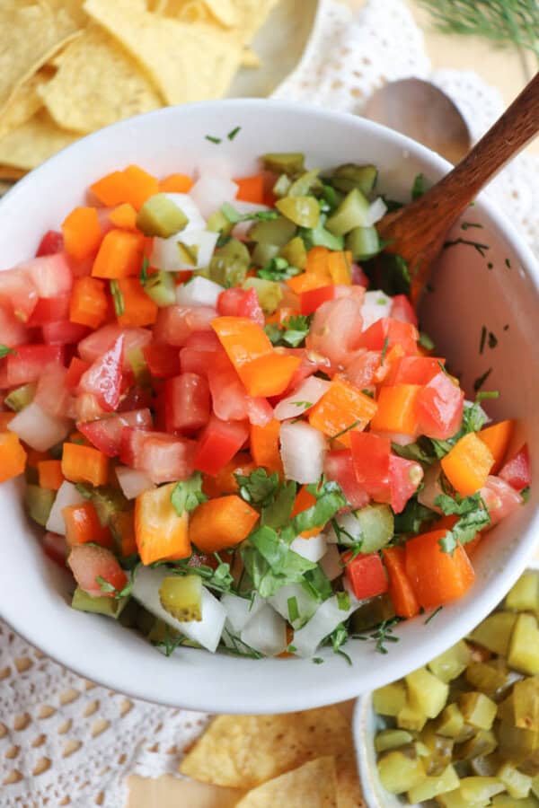 A bowl of colorful chopped vegetables, including tomatoes, orange bell peppers, onions, pickles, and cilantro, with a wooden spoon. Tortilla chips are visible in the background.
