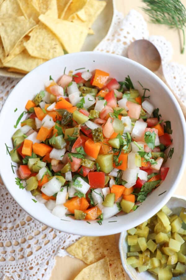 A white bowl filled with colorful chopped salad including tomatoes, onions, pickles, carrots, and herbs, placed on a lace doily with tortilla chips and a small dish of chopped pickles nearby.