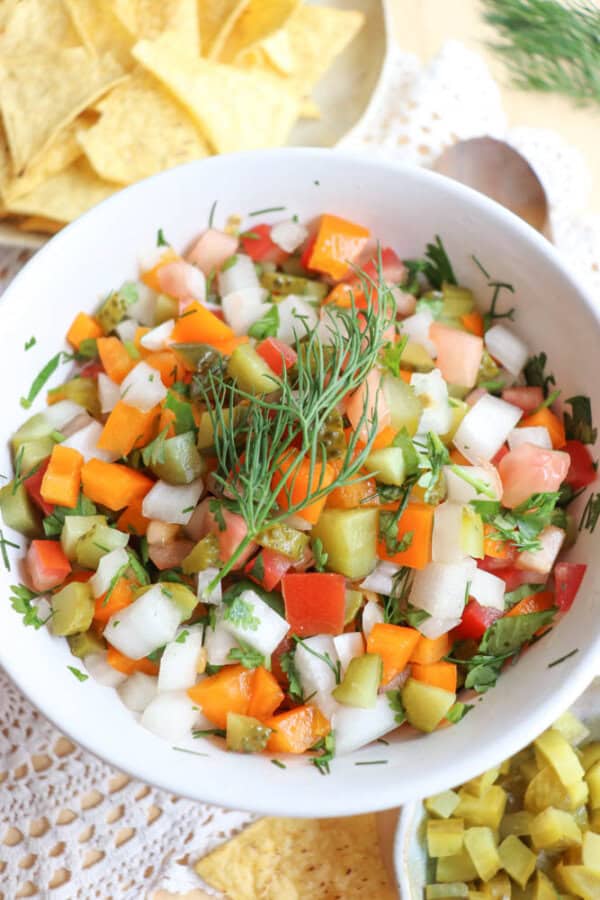 A white bowl filled with a colorful diced vegetable salad, including tomatoes, carrots, cucumbers, onions, and fresh herbs, garnished with a sprig of dill. Tortilla chips and a wooden spoon are nearby on a lace tablecloth.