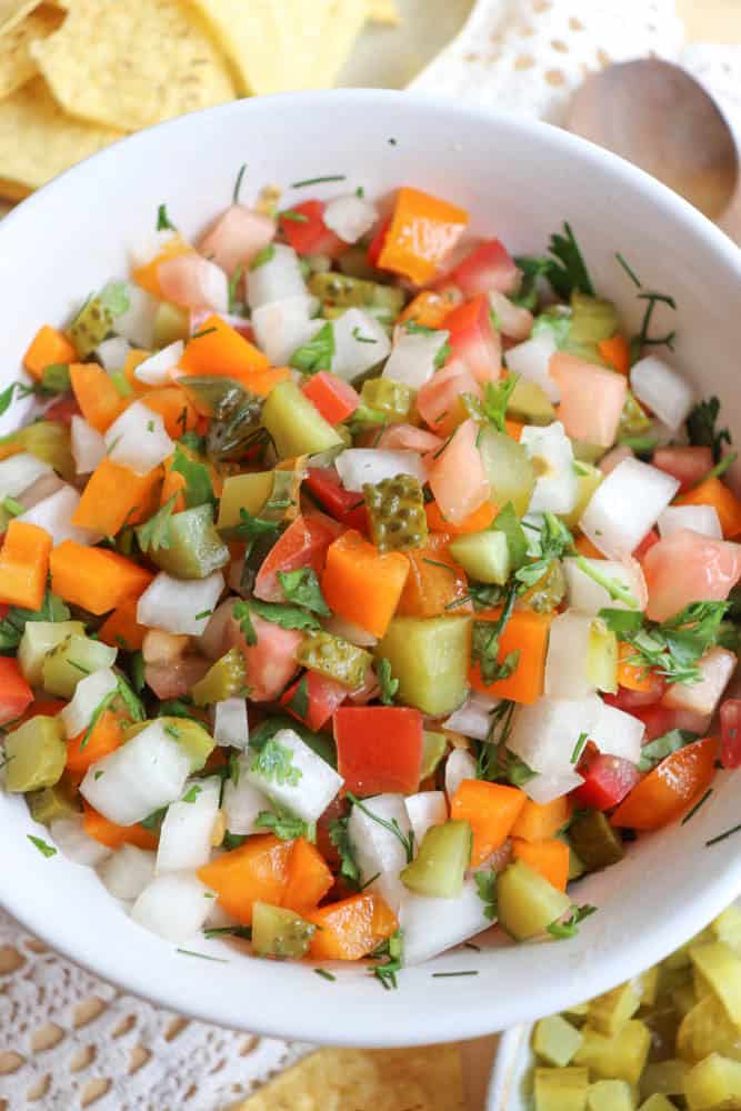A white bowl filled with colorful chopped vegetables, including tomatoes, onions, bell peppers, pickles, and fresh herbs, placed on a lace tablecloth. Tortilla chips are partially visible in the background.