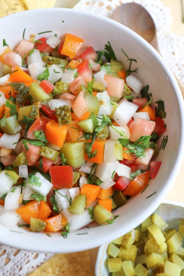 A close-up of a bowl filled with colorful chopped vegetables, including red and orange bell peppers, onions, tomatoes, pickles, and fresh herbs, creating a vibrant and fresh salsa or salad.