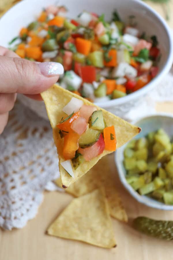 A hand holds a tortilla chip topped with colorful diced vegetables, including cucumbers, tomatoes, onions, and peppers, above a bowl of fresh salsa on a wooden table.
