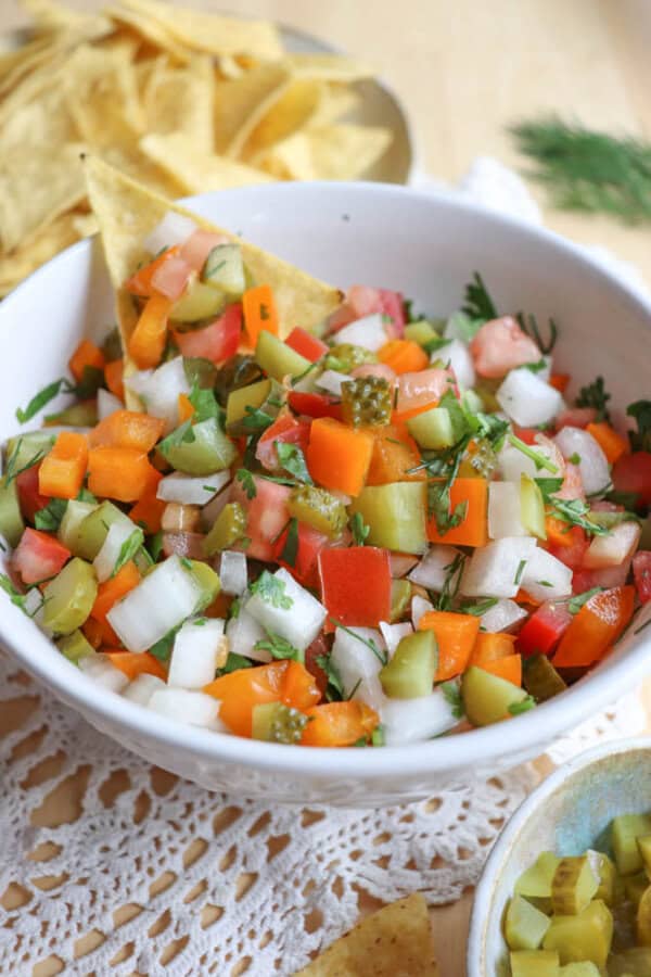 A bowl of colorful salsa with diced tomatoes, onions, pickles, bell peppers, and herbs, served with tortilla chips on a white lace tablecloth. A tortilla chip is dipped into the salsa.