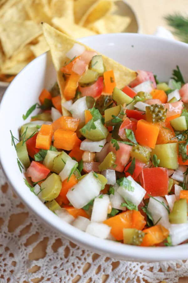 A white bowl filled with a colorful mix of chopped vegetables including tomatoes, onions, pickles, and bell peppers, garnished with herbs. A tortilla chip is dipped into the mixture, with more chips in the background.