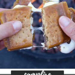 Close-up of hands pulling apart a gooey s’mores crack recipe treat, with melted marshmallow and chocolate stretching between two graham cracker bars. A fire pit with glowing coals is blurred in the background.