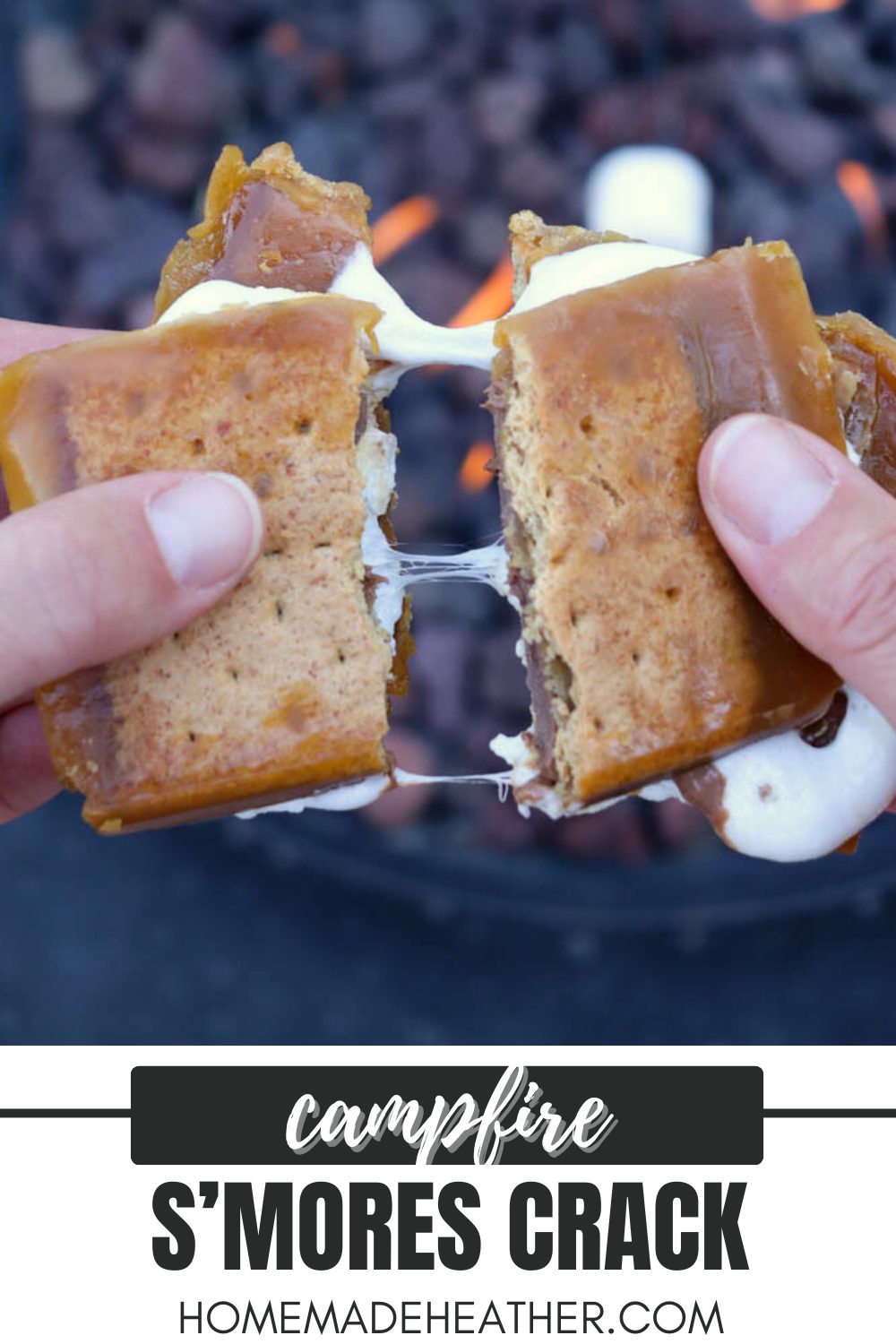 Close-up of hands pulling apart a gooey s’mores crack recipe treat, with melted marshmallow and chocolate stretching between two graham cracker bars. A fire pit with glowing coals is blurred in the background.