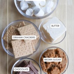 Five clear bowls on a wooden surface contain the essential smores crack recipe ingredients: marshmallows, graham crackers, butter, brown sugar, and chocolate. Each bowl is labeled, and the ingredients are neatly arranged in an overhead view.