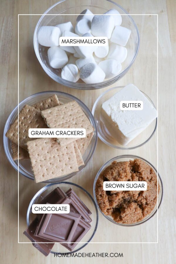 Five clear bowls on a wooden surface contain the essential smores crack recipe ingredients: marshmallows, graham crackers, butter, brown sugar, and chocolate. Each bowl is labeled, and the ingredients are neatly arranged in an overhead view.