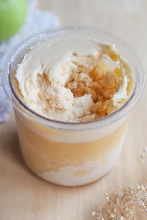 A close-up of a clear plastic cup filled with creamy, swirled vanilla and light caramel-colored ice cream or mousse, resting on a light wooden surface.