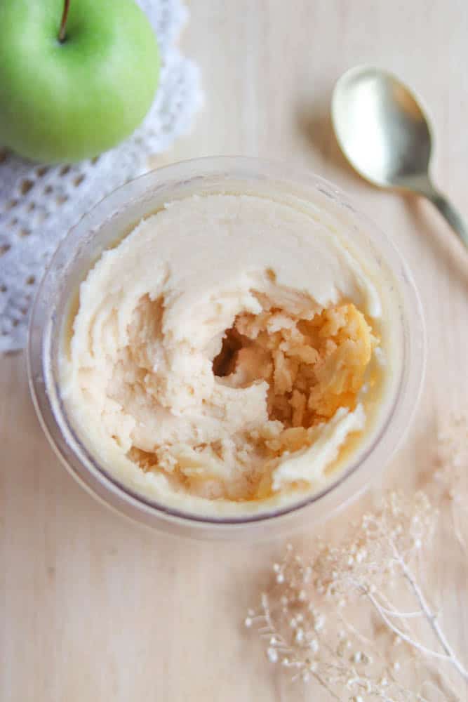 A glass jar filled with creamy apple butter is shown on a wooden surface, next to a gold spoon, a green apple, a white crocheted cloth, and some dried flowers.