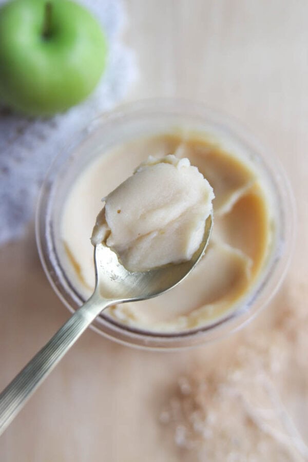 A spoon holding a scoop of smooth, creamy apple butter above a glass jar, with a green apple and lace cloth in the background.