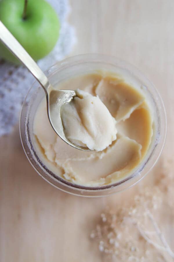 A spoon scooping creamy, beige apple butter from a small glass jar on a light wooden surface, with a green apple and lace cloth partially visible in the background.