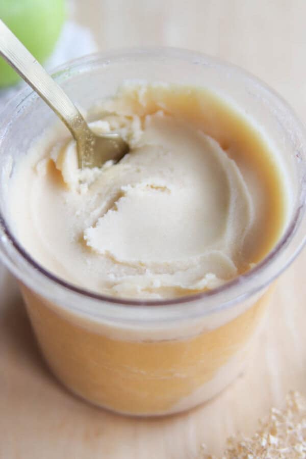 A close-up of a glass jar filled with creamy, light tan apple butter. A gold spoon is inserted into the jar, and the apple butter has a smooth, swirled texture.