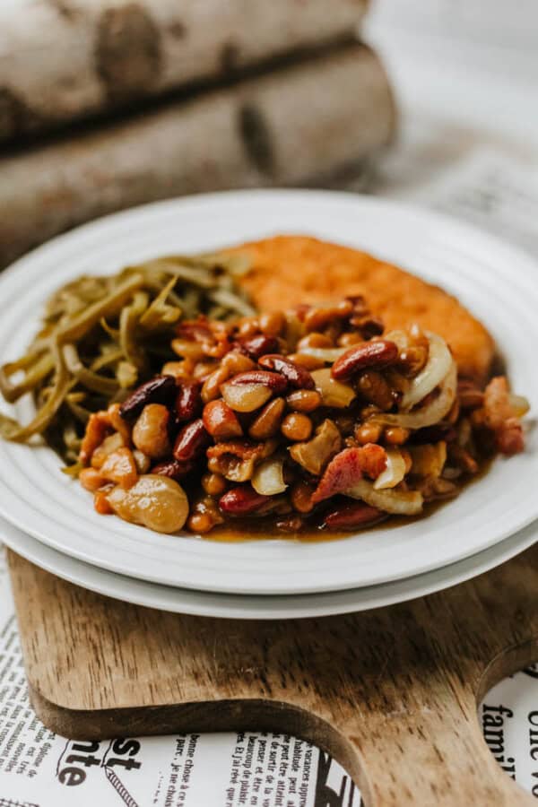 A white plate with baked beans, kidney beans, and onions, served with green beans and a breaded patty, sits on a wooden board over printed paper.
