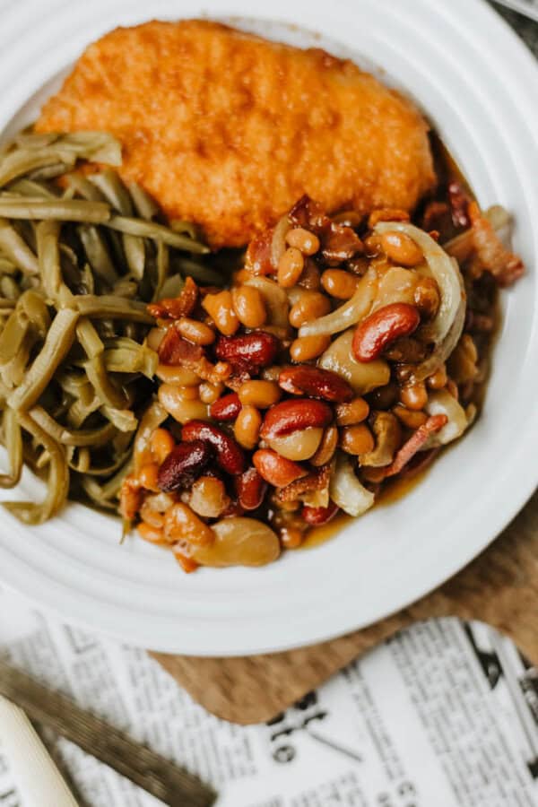 A plate with baked beans mixed with onions and bacon, cooked green beans, and a slice of cornbread, all served on a white dish.