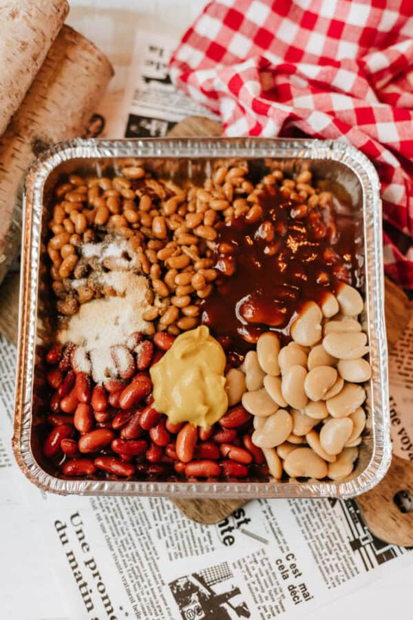 A foil tray filled with kidney beans, pinto beans, lima beans, barbecue sauce, mustard, and seasonings sits on a table covered with newspaper and a red-checkered cloth.