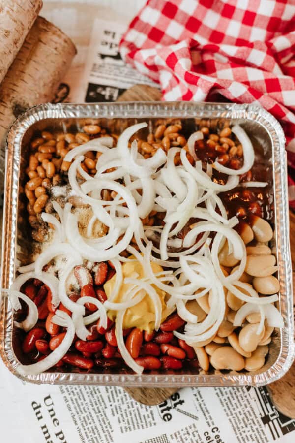 A foil tray filled with baked beans, kidney beans, lima beans, mustard, and sliced raw onions sits on newspaper with a red and white checkered cloth in the background.