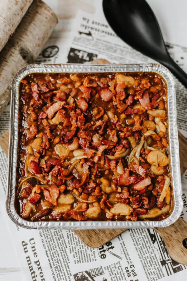 A square aluminum tray filled with baked beans topped with crispy bacon pieces and onions, placed on newspaper and a wooden board, with a black serving spoon nearby.