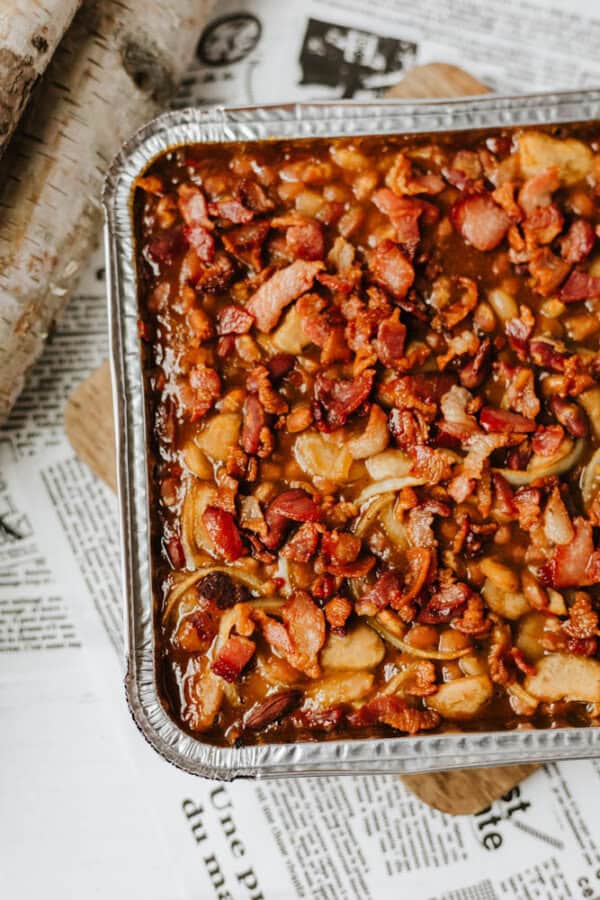 A close-up of a foil baking tray filled with a casserole topped with crispy bacon pieces and sliced potatoes, set on a table covered with newspaper.