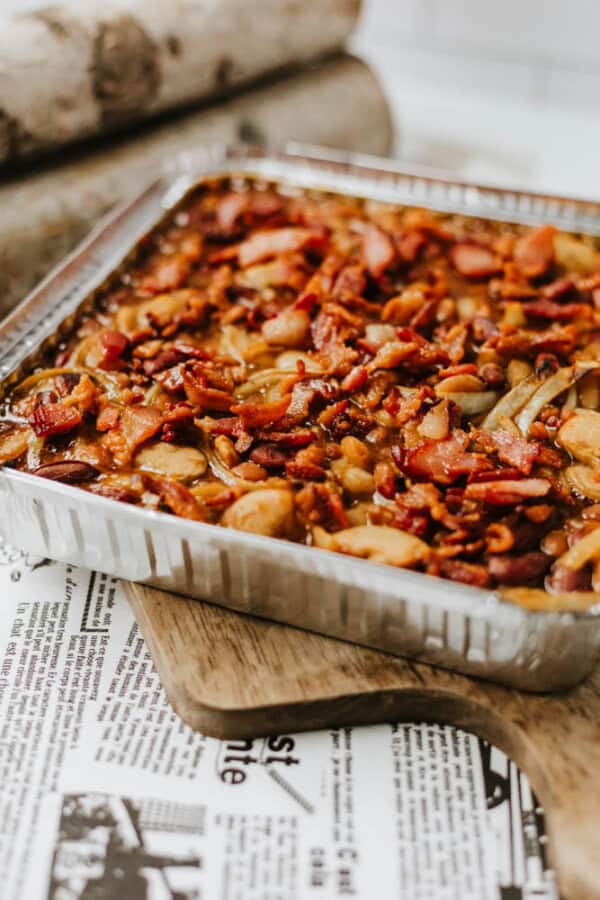 A foil tray filled with baked beans, sliced sausages, and crispy bacon pieces sits on a wooden board covered with newspaper print.