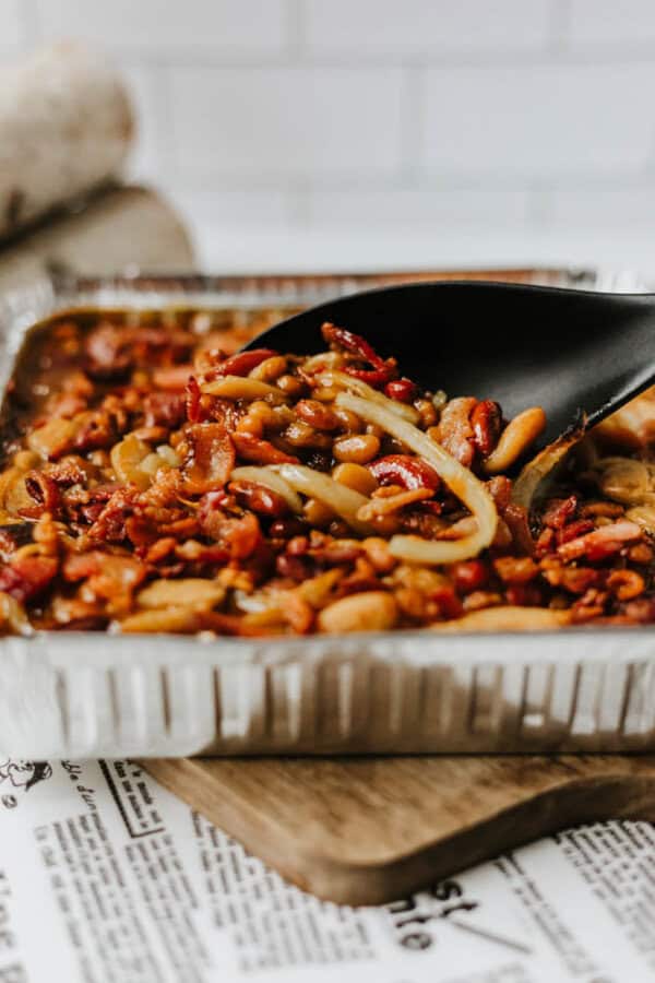 A close-up of a black spoon scooping a baked bean casserole with onions and bacon from a foil tray, resting on a wooden cutting board.