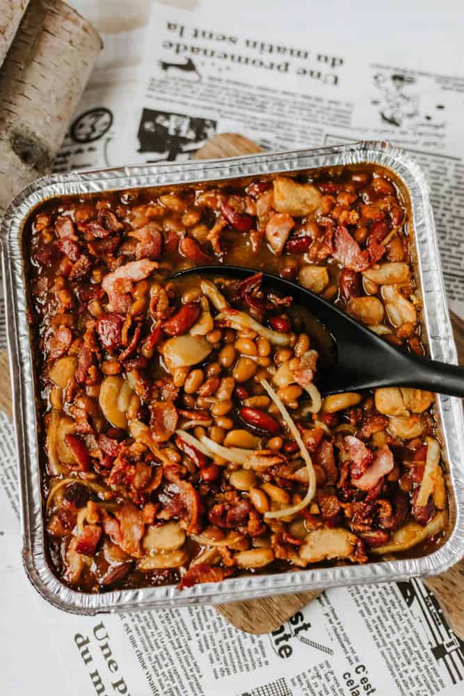 A foil tray filled with baked beans, bacon pieces, and mixed beans, with a black serving spoon resting inside. The tray sits on newspaper pages.