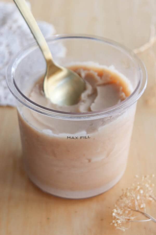 A clear plastic cup filled with a creamy beige dessert is shown on a wooden surface. A gold spoon rests inside, and “MAX FILL” is printed on the cup. Dried flowers and a white lace cloth are in the background.