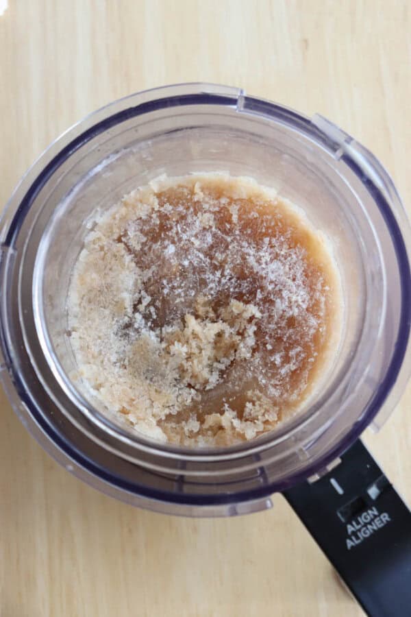 A top-down view of a food processor bowl containing a mixture of brown and white granulated ingredients on a light wooden surface.