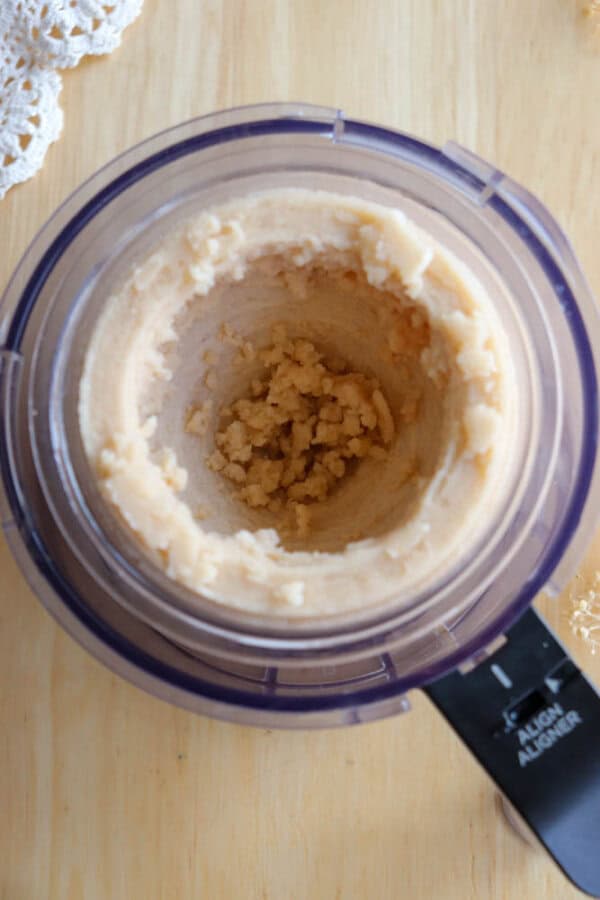 A food processor with a creamy, light beige mixture partially blended inside, viewed from above, on a wooden surface with a bit of lace cloth visible in the corner.