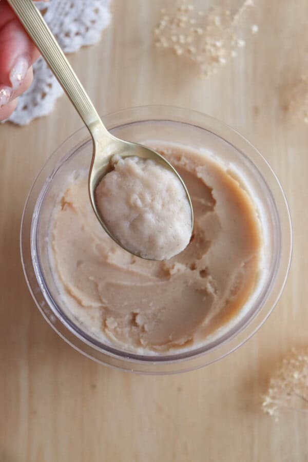 A hand holds a spoon scooping a creamy beige substance from a clear container on a wooden surface, with lace and granules scattered nearby.