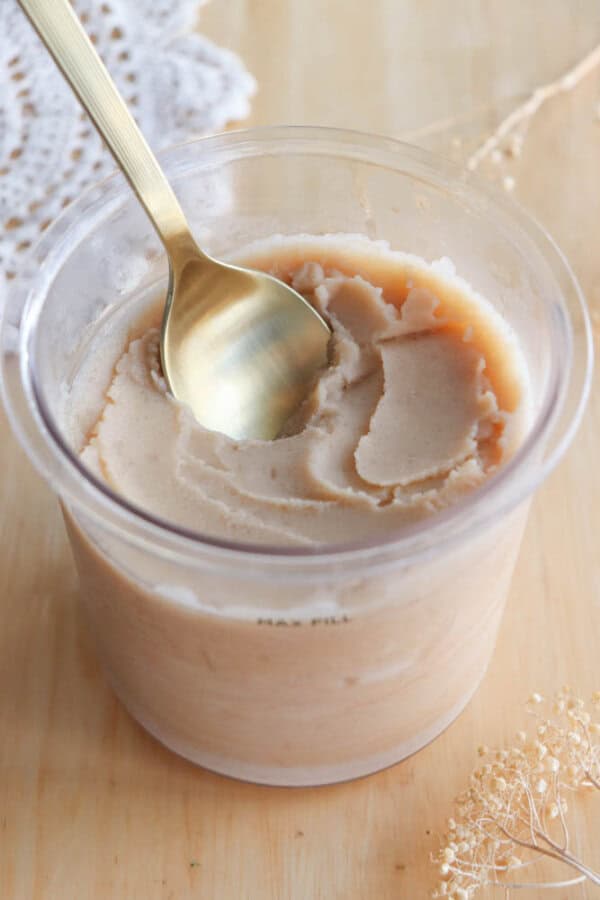 A clear plastic container filled with smooth, light brown nut butter. A gold spoon is scooping some of the spread, and dried flowers are visible on the wooden surface beside the container.