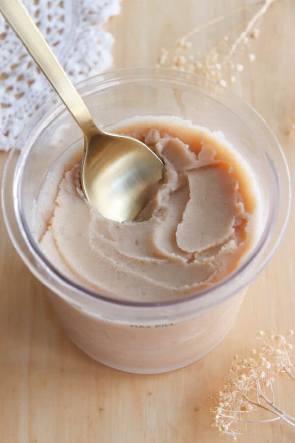 A gold spoon scoops into a container of light brown, creamy ice cream on a wooden surface, with delicate dried flowers and a piece of lace in the background.