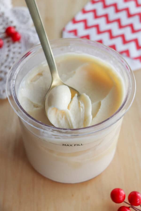 A spoon scooping creamy, white ice cream from a clear plastic container placed on a light wooden surface, with red berries and a red and white patterned cloth nearby.