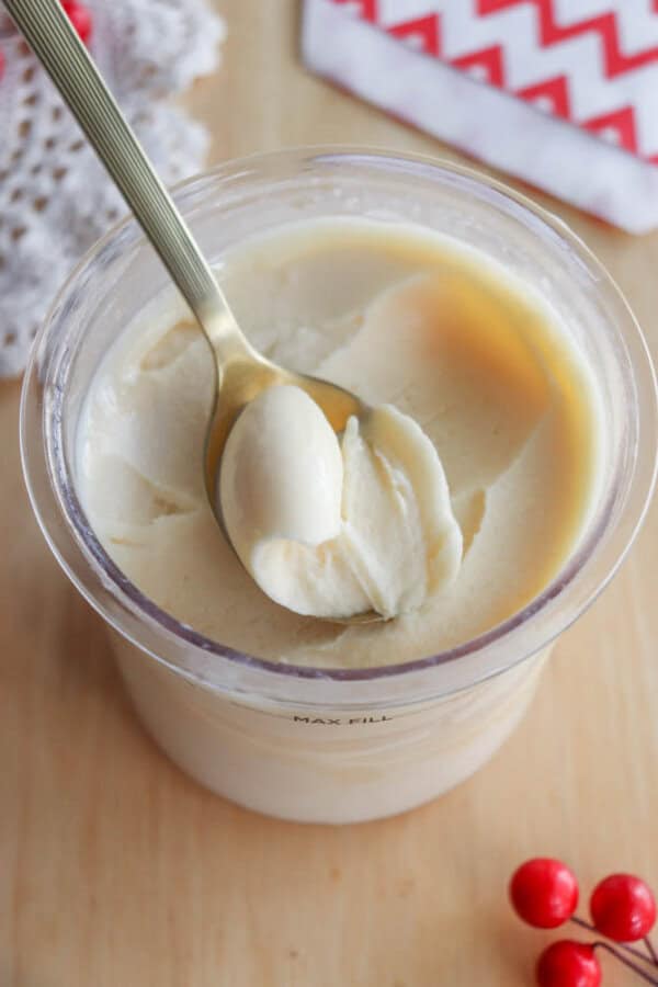 A spoon is scooping creamy, white ice cream or frozen dessert from a clear plastic container on a wooden surface. Red berries and a white doily are nearby, with a red and white napkin in the background.