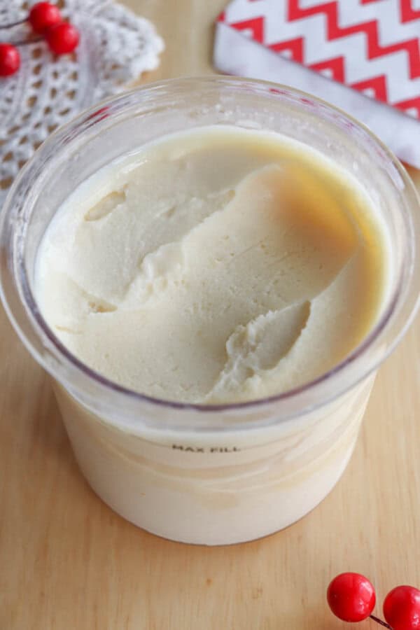 A container filled with creamy vanilla ice cream sits on a wooden surface, surrounded by a doily, red berries, and a red-and-white chevron-patterned napkin.