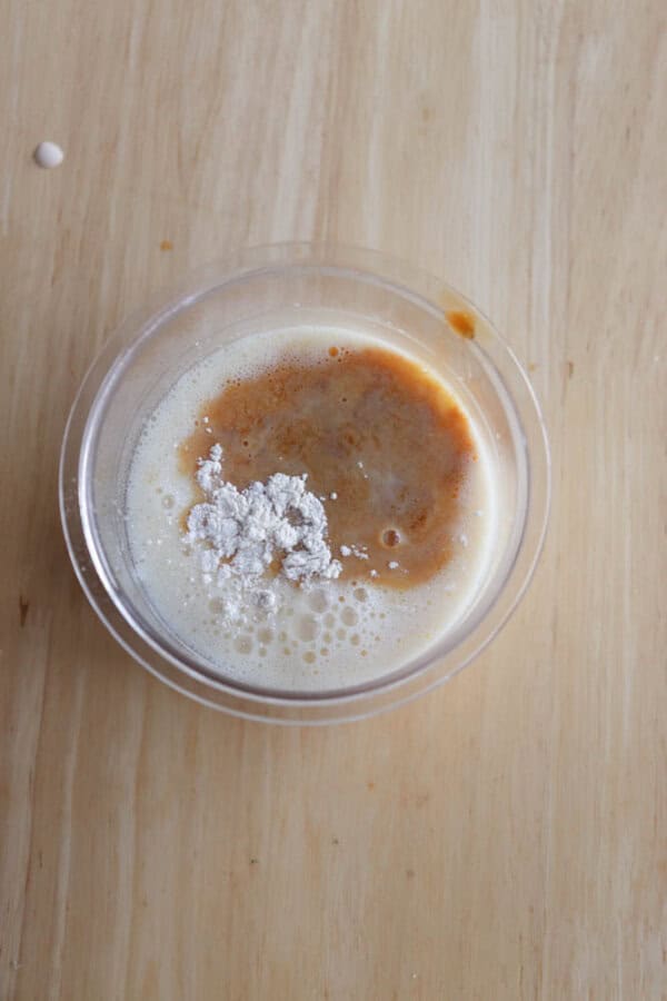 A clear bowl contains a foamy liquid mixture topped with a brown liquid and a small mound of white powder, sitting on a light wooden surface.