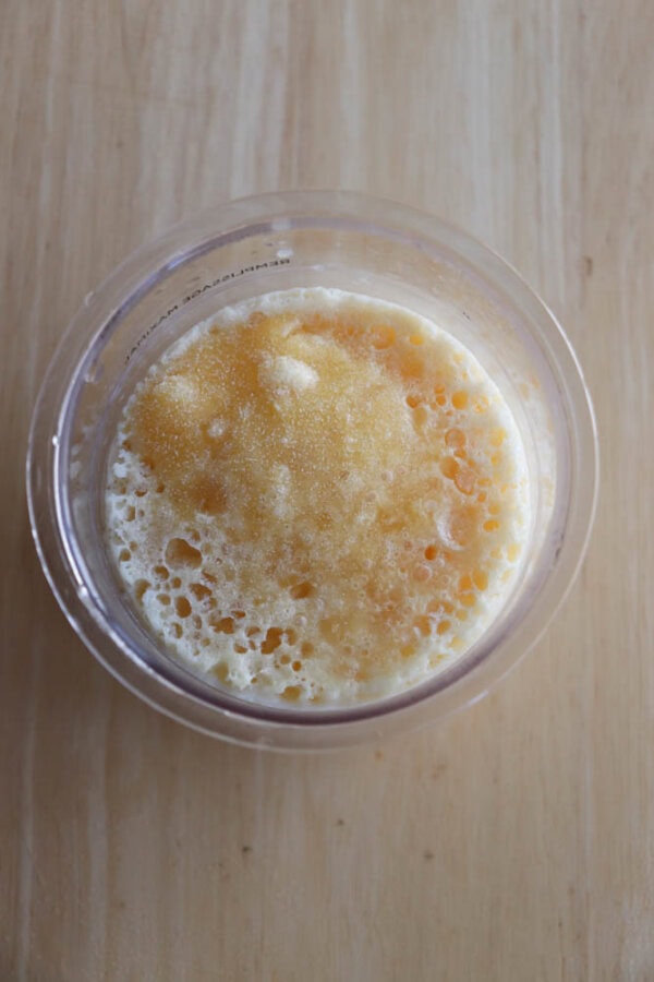 A clear cup filled with a mixture of milk and granulated sugar, with some sugar floating on top and small bubbles around the edges, sits on a light wooden surface.