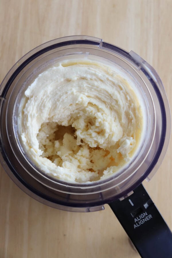 A food processor bowl filled with blended and creamy yellow dough or batter, sitting on a light wooden surface.