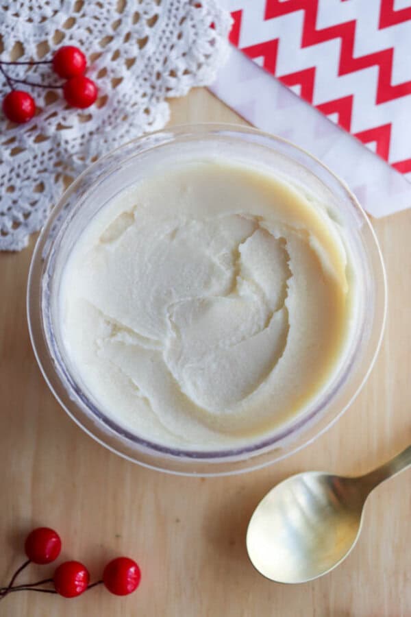 A glass container filled with creamy white body butter sits on a wooden surface beside a gold spoon, red berries, a lace doily, and a red-and-white chevron-patterned napkin.