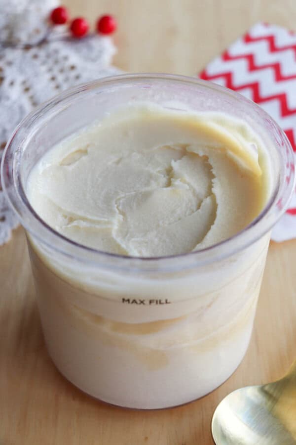 A close-up of a clear container filled with creamy, off-white butter or spread, labeled MAX FILL. The background shows a lace cloth, a red-and-white chevron napkin, and part of a gold spoon.