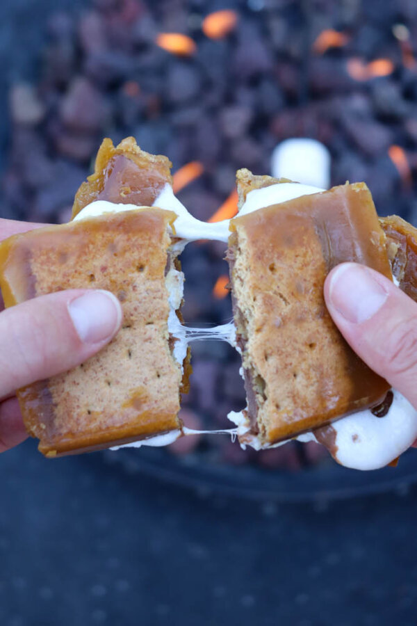 Two hands pull apart a gooey s’more made with golden brown cookies, melted chocolate, and marshmallow—just like a homemade smores crack recipe—with a fire pit burning in the blurred background.
