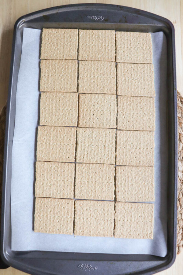 A baking sheet lined with parchment paper holds 18 rectangular graham cracker sheets arranged neatly in rows, the perfect base for any smores crack recipe.